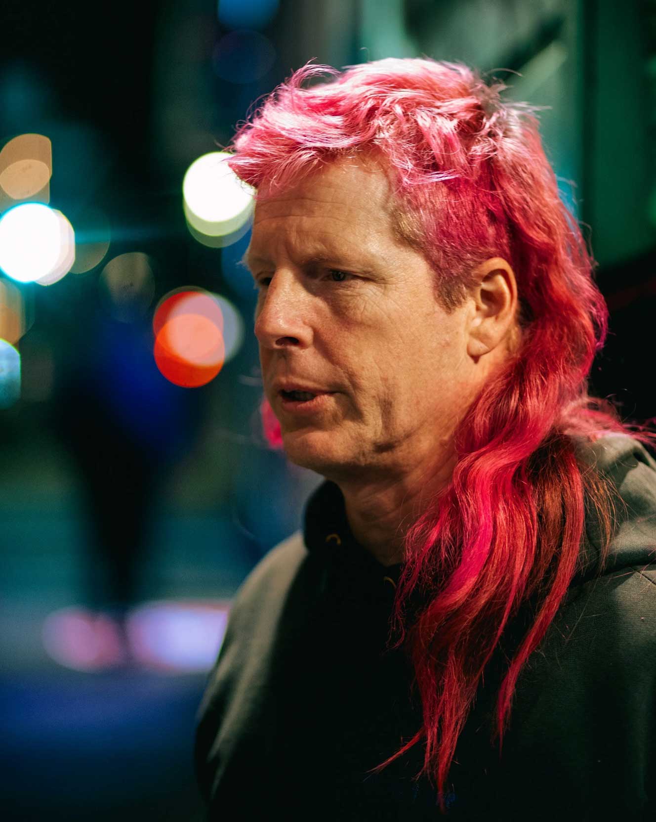 Color portrait of Chris Moore with a serious expression sporting a long pink mullet and a black hoodie